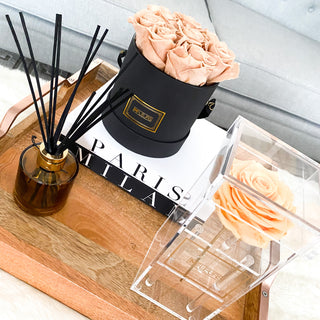 A photo setup with black round rose box and a Solitaire rose box with peach colored roses on a wooden tray with black and white books and sented sticks.